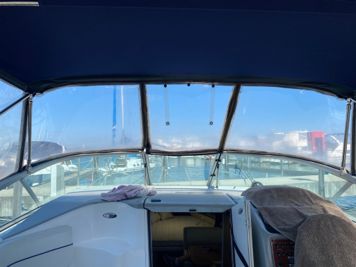 Interior view of a boat's cockpit with a clear windshield and protective clears overlooking the marina, featuring a control panel, white seating, and a blue canopy above.