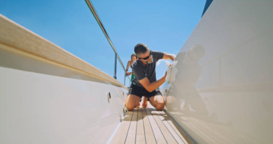 Man in sunglasses kneeling and cleaning the wooden deck of a luxury yacht under bright sunlight, with a clear blue sky overhead.