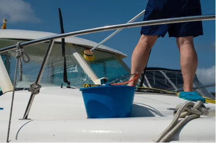 Person cleaning a yacht's windshield with a brush and soapy water, with a blue bucket and ropes on the deck under a clear sky.