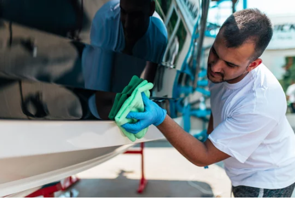Man in a white shirt and blue gloves meticulously cleaning the side of a boat with a green cloth, with a reflection visible on the boat's surface.